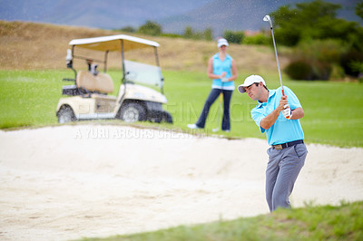 Buy stock photo A young male golfer chipping his ball out of a bunker while his female partner looks on from the fairway