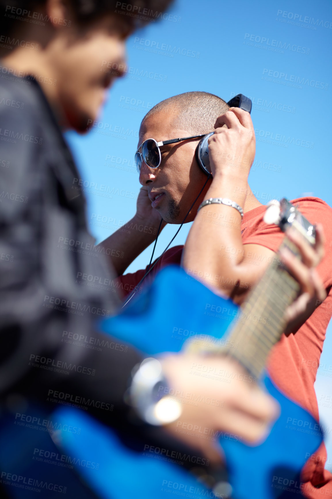 Buy stock photo Shot of a young man playing the guitar while his friend uses his his headphones