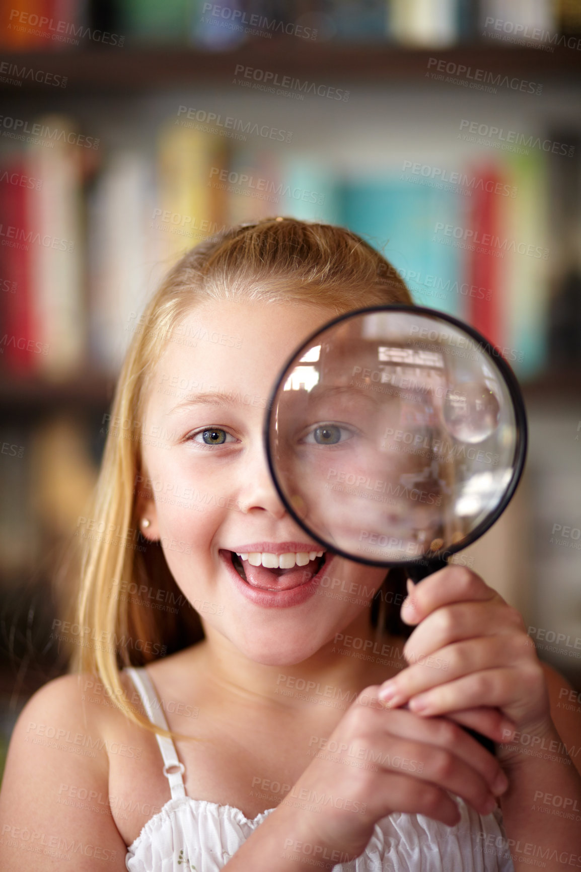 Buy stock photo Portrait of excited curious little girl looking through magnifier while in front of a book shelf in a library. Adorable inquisitive little school kid using magnifying glass and expressing amazement 