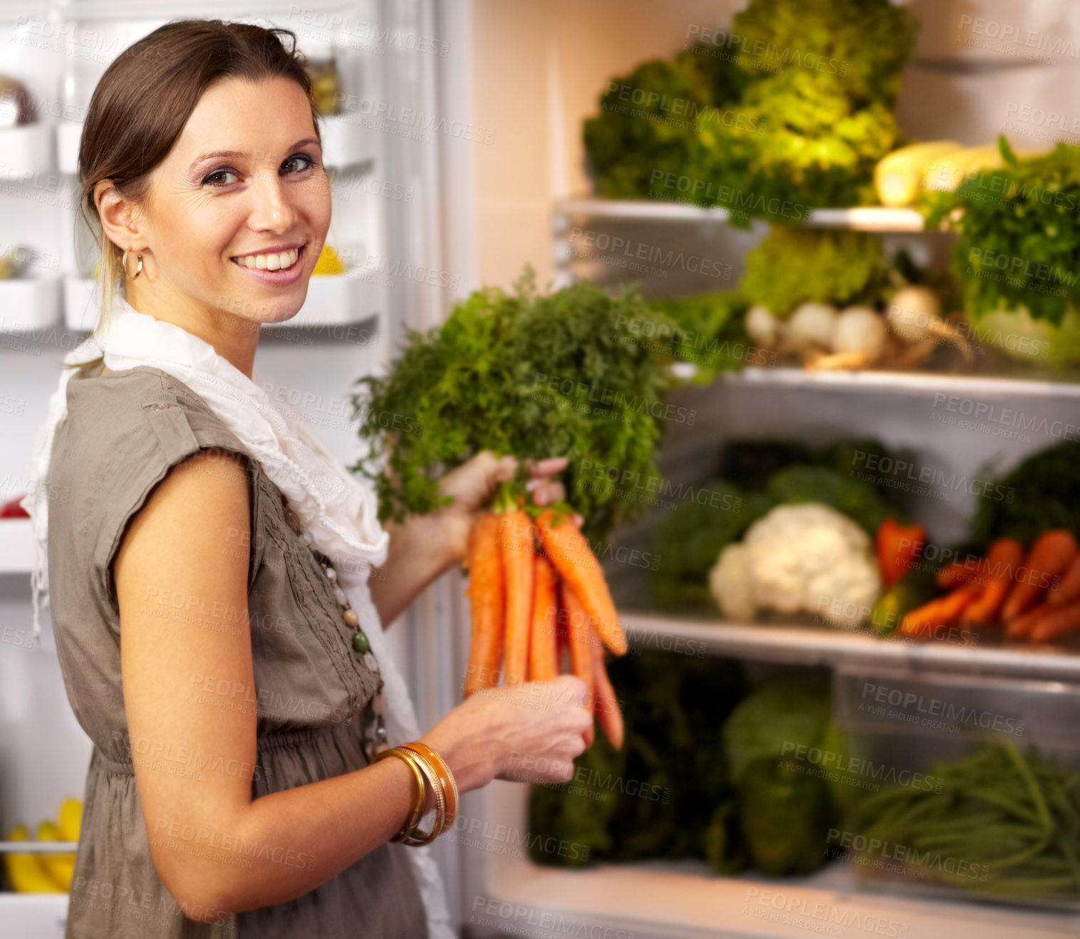 Buy stock photo Smiling woman adding some fresh carrots to her fridge already stocked with raw vegetables