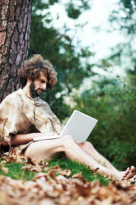 Buy stock photo A confused looking caveman sitting by a tree with a laptop