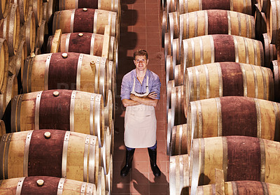 Buy stock photo Portrait of a young male winemaker standing with his arms crossed next to wooden barrels of red wine in a cellar at a distillery. Entrepreneur built his business to the point of success