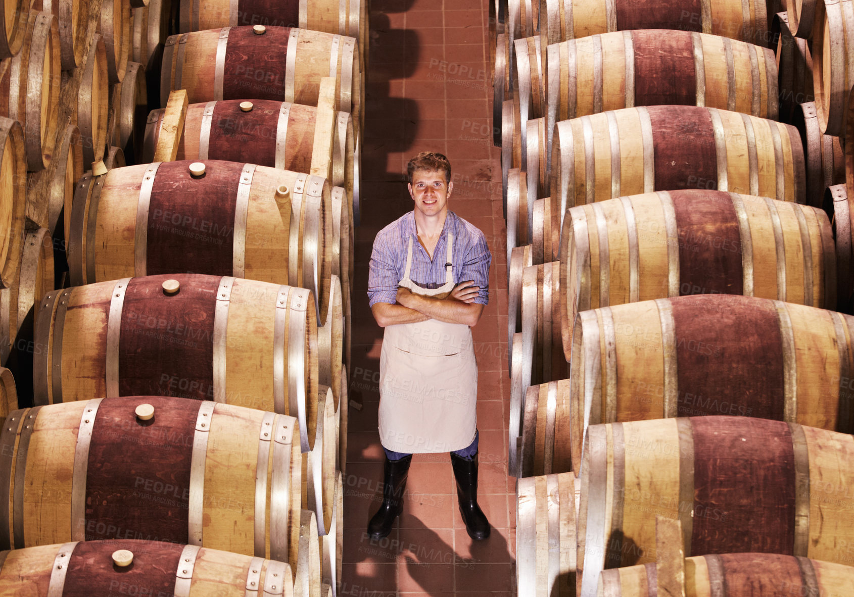 Buy stock photo Portrait of a young male winemaker standing with his arms crossed next to wooden barrels of red wine in a cellar at a distillery. Entrepreneur built his business to the point of success