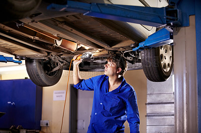 Buy stock photo A young male mechanic using a light to look underneath a car