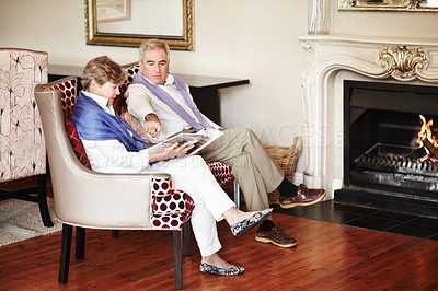 Buy stock photo Senior couple, relax and reading by fire place on holiday, retirement or vacation together at hotel or resort. Man and woman sitting with magazine or paper for news, deals or activities at house