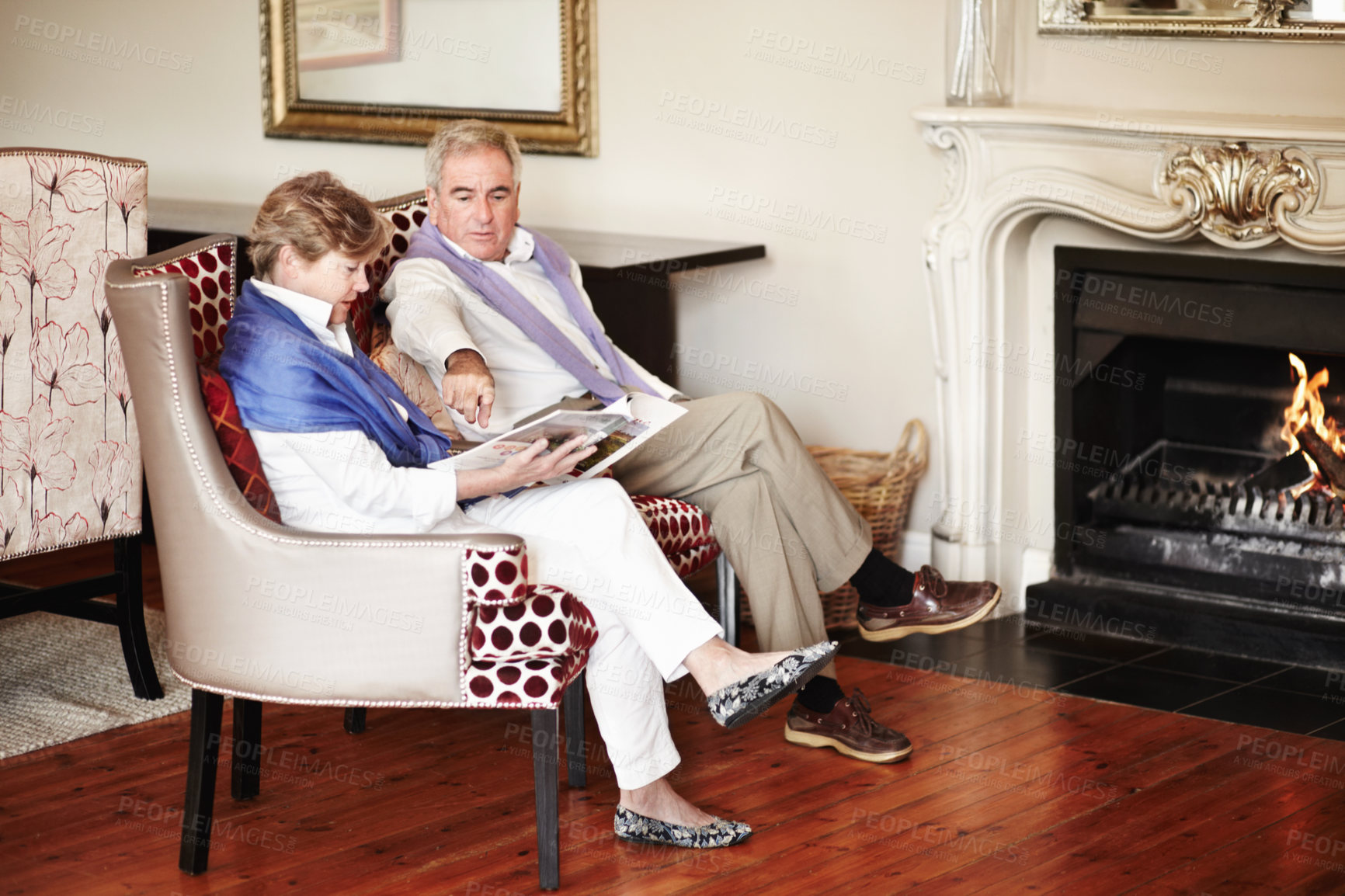 Buy stock photo Senior couple, relax and reading by fire place on holiday, retirement or vacation together at hotel or resort. Man and woman sitting with magazine or paper for news, deals or activities at house