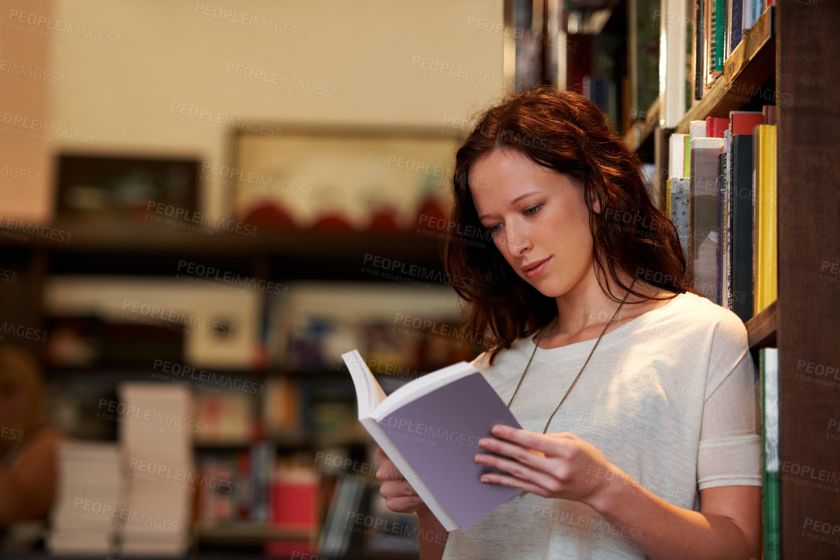 Buy stock photo A young woman lost in the pages of a novel against a backdrop of full bookshelves