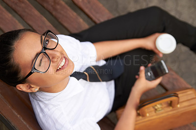 Buy stock photo A beautiful young woman winking at the camera while sitting on a bench with her cellphone, coffee and satchel