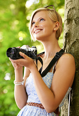 Buy stock photo Cute young photographer holding her camera and leaning against a tree
