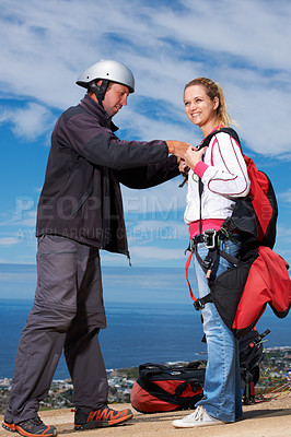 Buy stock photo Man, woman and ready with safety for paragliding, extreme sport and helping by ocean in sunshine. Coaching, fitness and adventure with helmet, backpack and parachute for jump, outdoor and blue sky