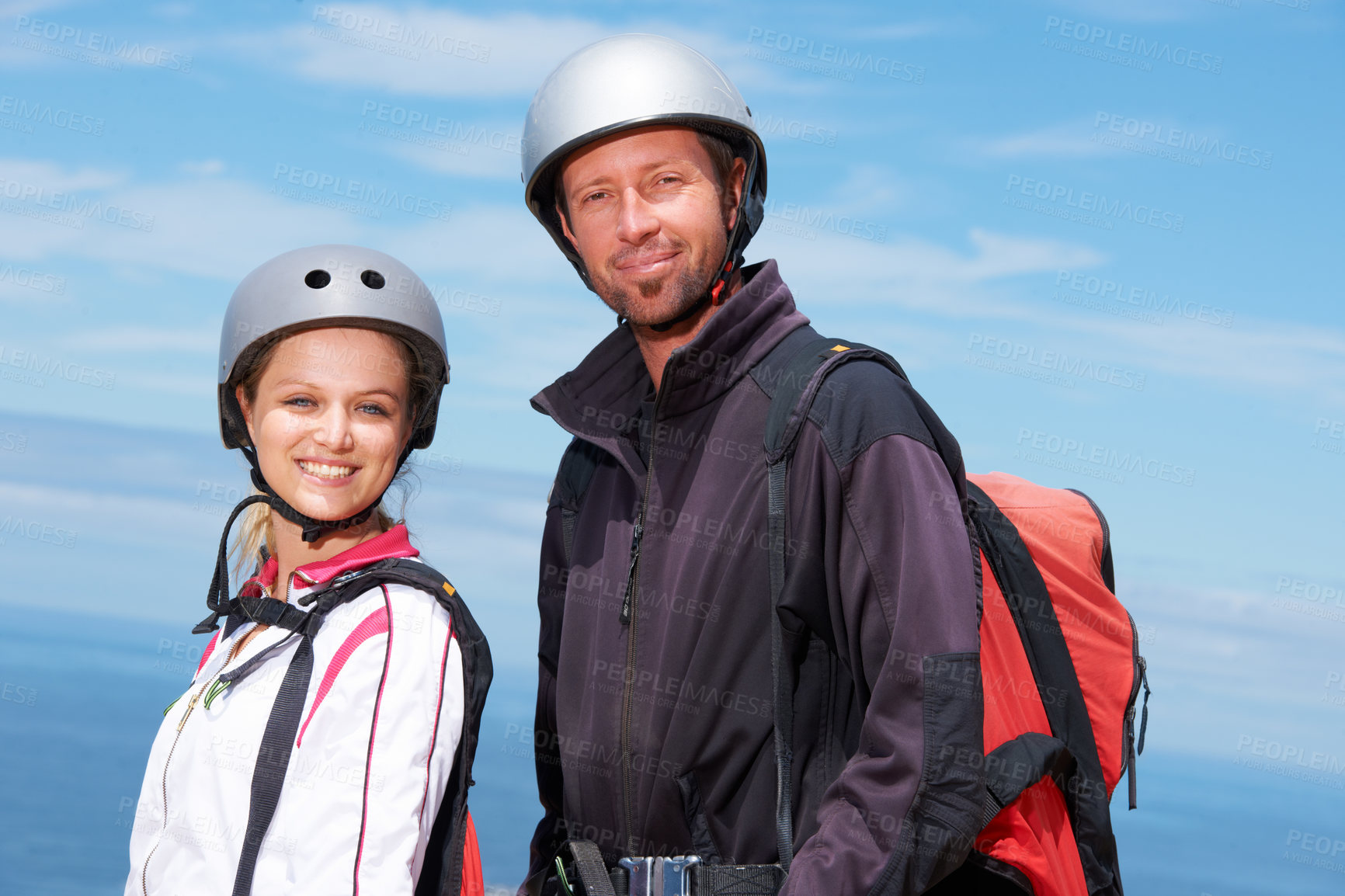 Buy stock photo Man, woman and portrait with smile for paragliding, extreme sport and outdoor by sea in sunshine. Happy couple, fitness and adventure with helmet, backpack or parachute for jump, safety and blue sky