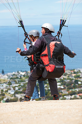 Buy stock photo People, paragliding and outdoor to land on ground, extreme sport and sunshine by ocean on hill. Coach, person and back on adventure with helmet, backpack or parachute for jump, safety and blue sky