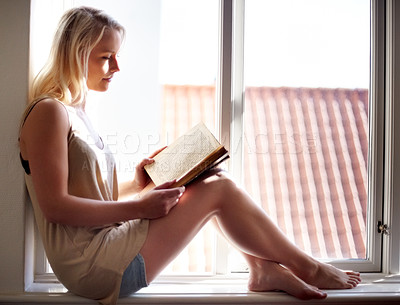 Buy stock photo A young woman relaxing on a window sill and reading an absorbing novel