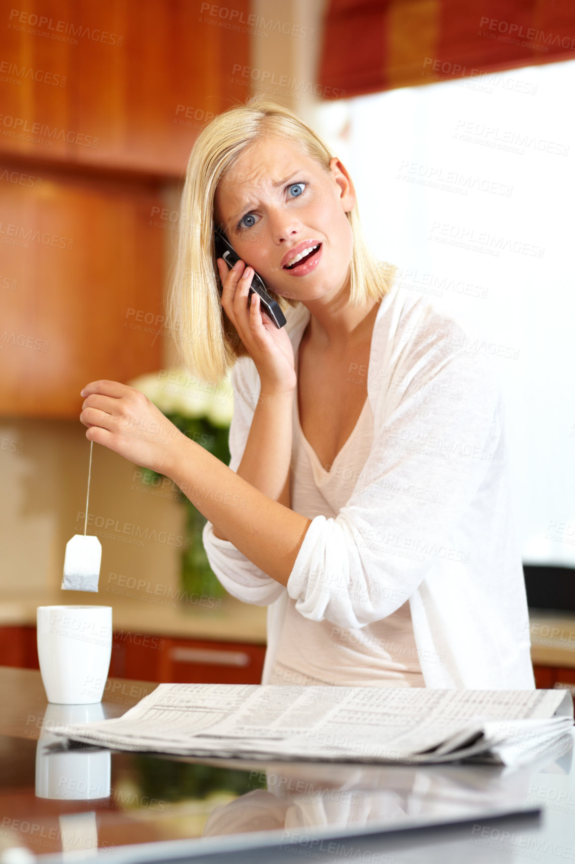 Buy stock photo A lovely young woman taking a phone call while making tea and reading the newspaper