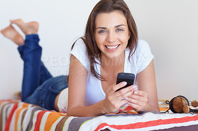 Buy stock photo Portrait of a smiling teenage girl relaxing on her bed texting on her phone with her headphones beside her