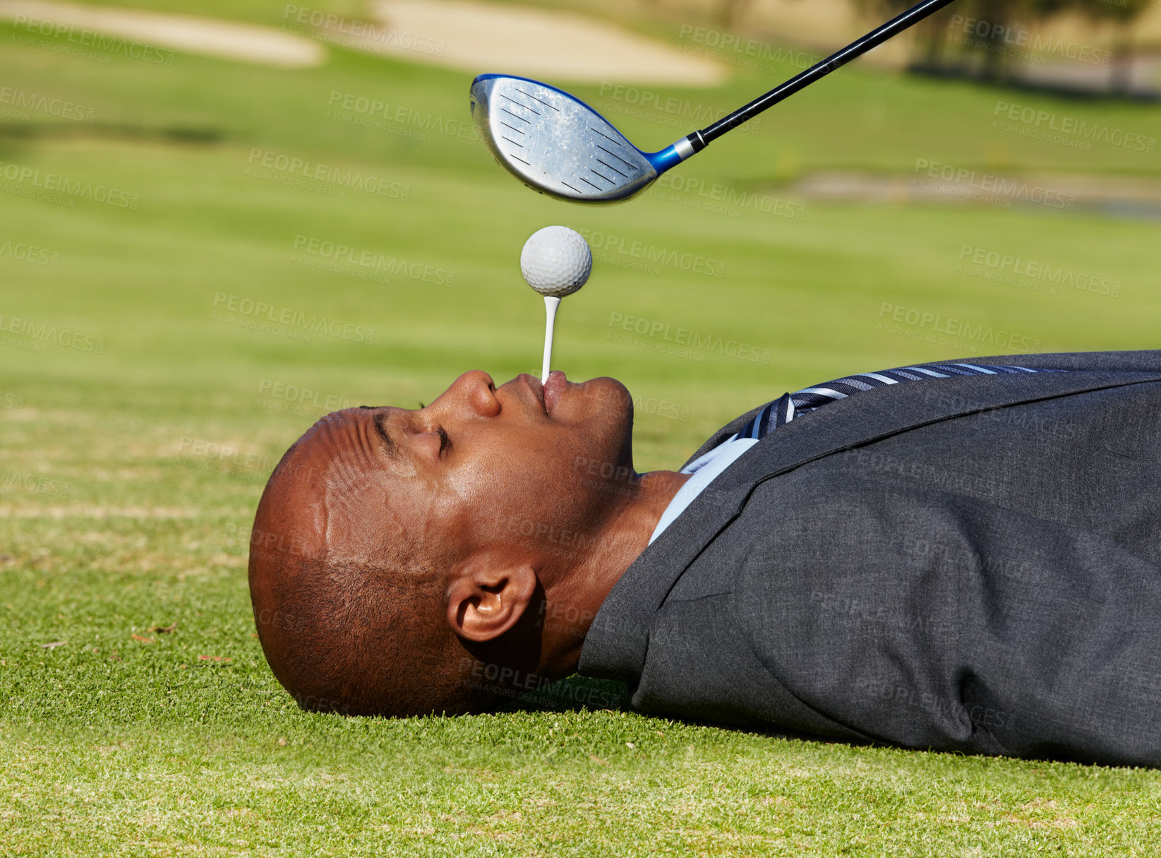 Buy stock photo Black man, face and lying on golf course with tee in mouth for hole in one on green grass or field. African male person, golfer or assistant holding ball with club hovering for swing or hit on pitch