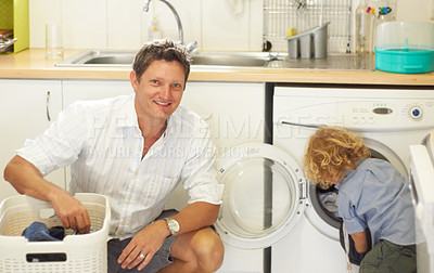 Buy stock photo Portrait of a father crouched next to a washing basket while his toddler sun fills up the washing machine with clothes