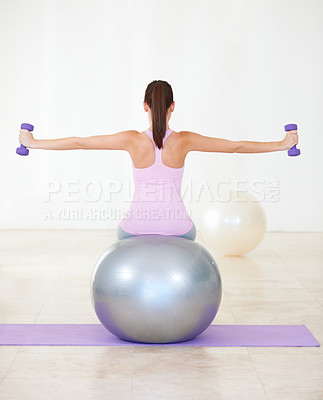 Buy stock photo Rearview shot of a young woman sitting on an exercise ball and doing some strengthening exercises with dumbbells
