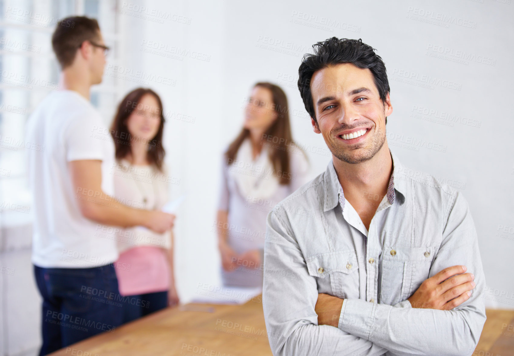 Buy stock photo Portrait of a young business professional standing with his arms folded with coworkers talking in the background