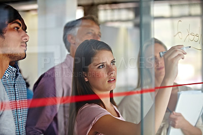 Buy stock photo A young businesswoman writing down plans on a glass pane while her associates look on