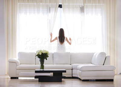 Buy stock photo A young woman looking through her living room window early in the morning