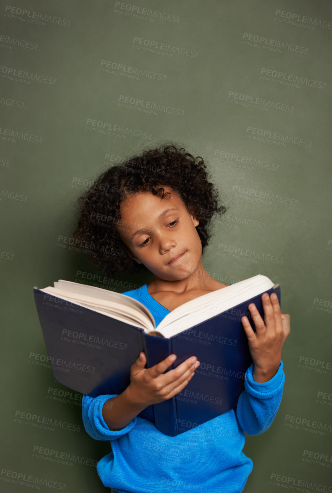 Buy stock photo A young ethnic boy reading a book while standing in front of a blackboard