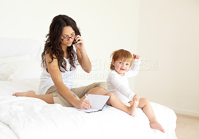 Buy stock photo A young mother speaking on her phone and writing in a notebook while her toddler sits with her on the bed