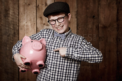Buy stock photo Young boy in retro clothing holding his piggybank while pointing towards it