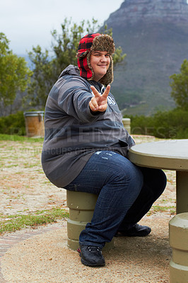 Buy stock photo An obese young man sitting outside at a picnic table showing a peace-sign