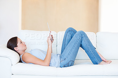 Buy stock photo A pretty young woman lying on the couch and browsing the internet on her tablet