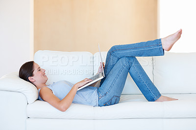 Buy stock photo A pretty young woman lying on the couch and browsing the internet on her laptop