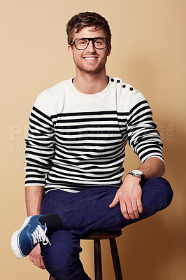 Buy stock photo Portrait, fashion and glasses with a man on a chair in studio on tan background for trendy style. Relax, casual and eyewear with a confident young geek or nerd sitting on a stool in a clothes outfit