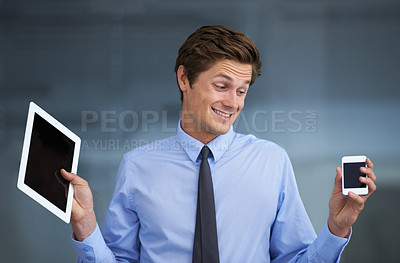 Buy stock photo A smiling young businessman trying to make a choice between a tablet or a smartphone
