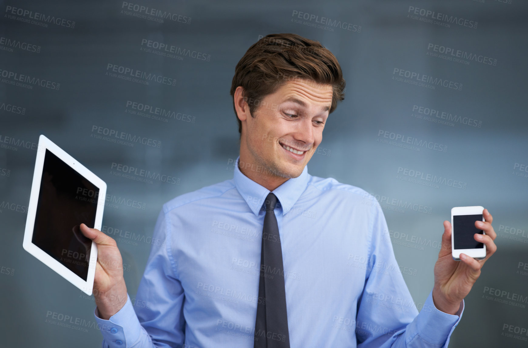 Buy stock photo A smiling young businessman trying to make a choice between a tablet or a smartphone