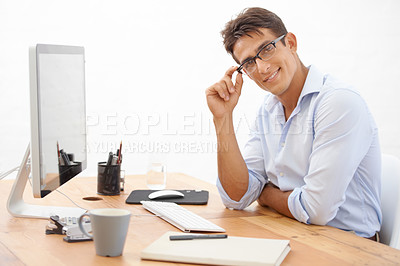 Buy stock photo A young businessman touching his glasses while smiling at the camera from behind his desk