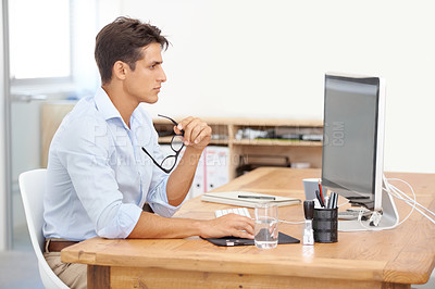 Buy stock photo A handsome young businessman holding his glasses while looking thoughtfully at his computer screen