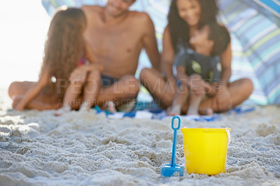 Buy stock photo Family, sand and relax on beach with bucket, spade and umbrella for playing by ocean coast. Blue and yellow toy on sandy shore with people in fun bonding, shade or outdoor holiday weekend together