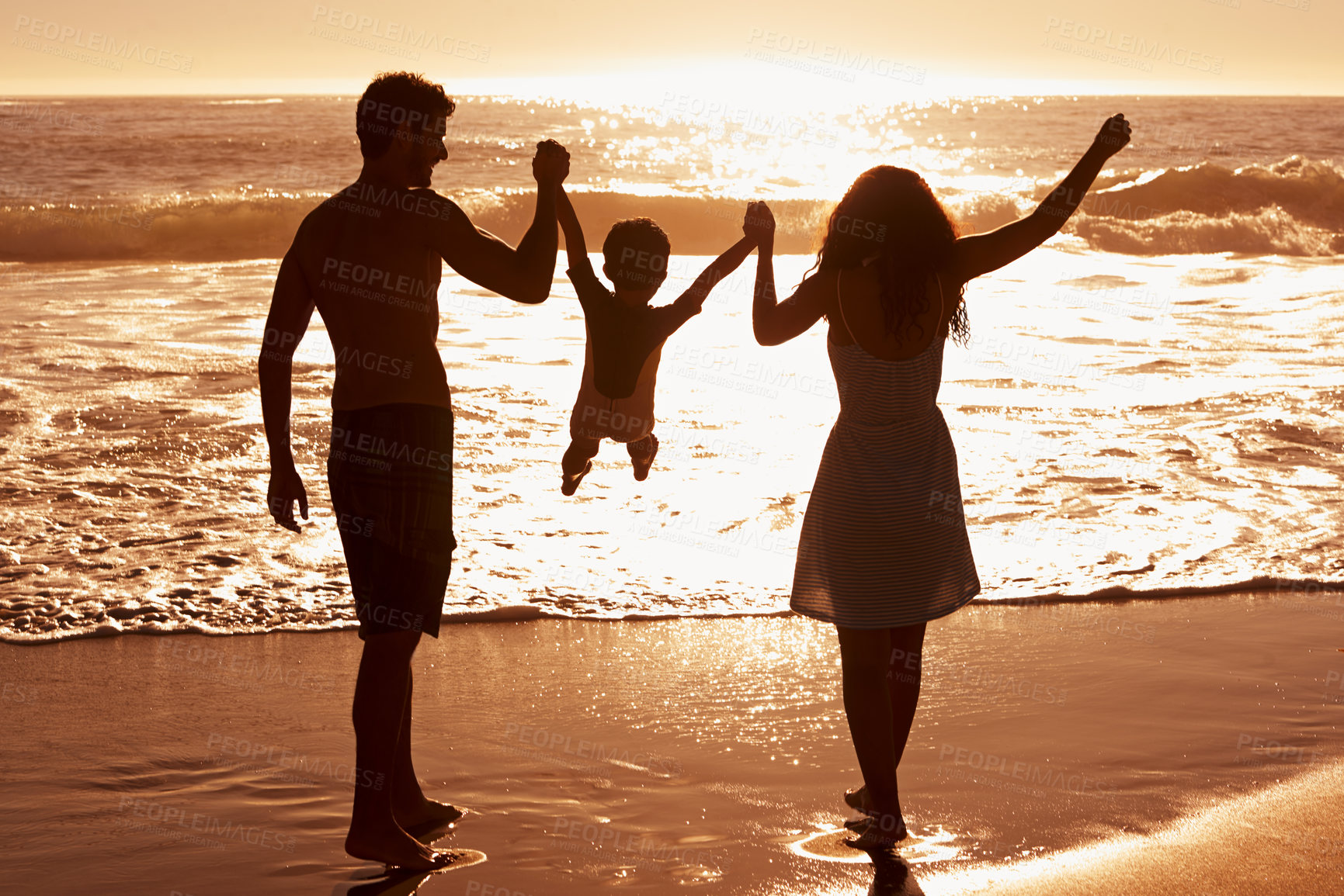 Buy stock photo Rear-view of a couple swinging their child between them as they walk on the beach at sunset