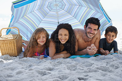 Buy stock photo A happy family smiling at the camera while lying under an umbrella at the beach