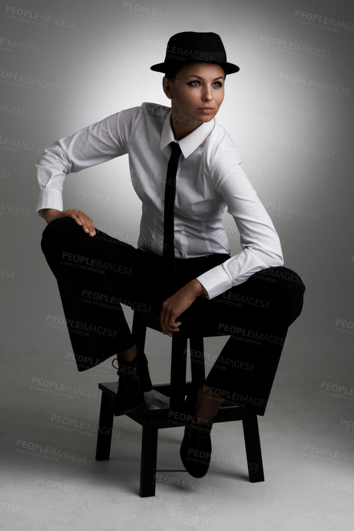 Buy stock photo A young woman wearing a shirt and tie and fedora posing on a stool against a grey background