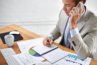 Buy stock photo Shot of a young businessman talking on the phone while writing at his desk