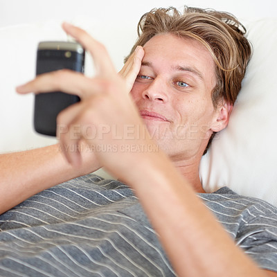 Buy stock photo Shot of a handsome young man smiling and holding a phone while lying down