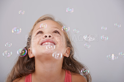 Buy stock photo Smile, little girl and bubbles in studio for fun, excited and enjoy games with soap liquid magic. Child, happy and learning to play with bubble, natural and wellness in looking up by gray background