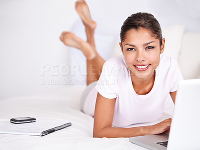 Buy stock photo Shot of an attractive young woman lying on her bed working on her laptop with her phone and a document next to her