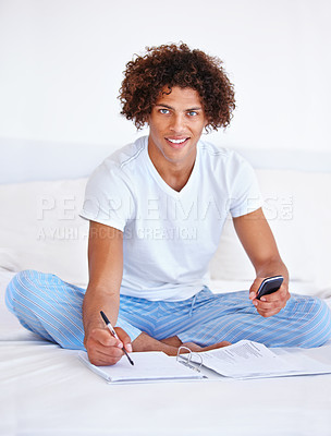 Buy stock photo Portrait of a handsome young man sitting in bed holding a phone and writing in a binder