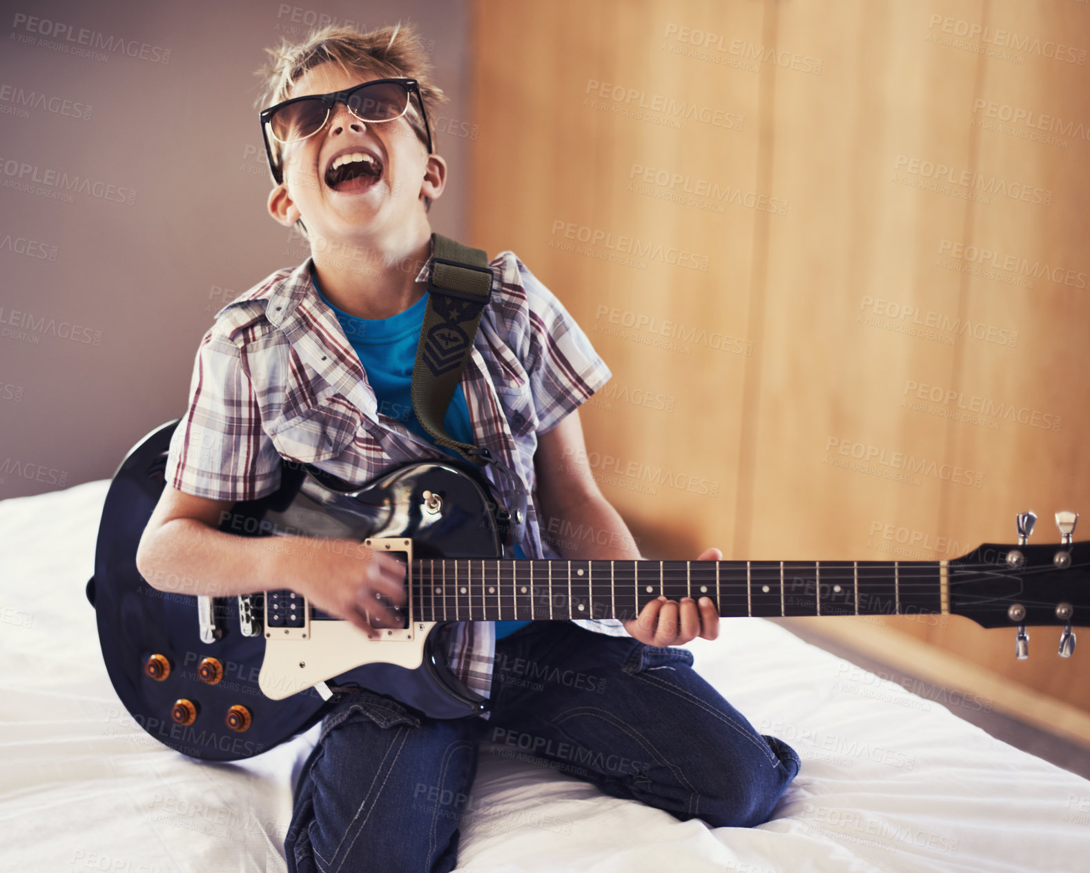 Buy stock photo Happy, playing and child with guitar on bed for music lesson with sunglasses at modern home. Fun, rockstar and young boy kid learning with electric string instrument in bedroom for hobby at house.