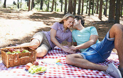 Buy stock photo Happy couple, kiss and relax in nature for picnic, love or support in affection, date or outdoor bonding. Woman and man sitting on floor with basket of fruit for embrace, eating or romance in forest