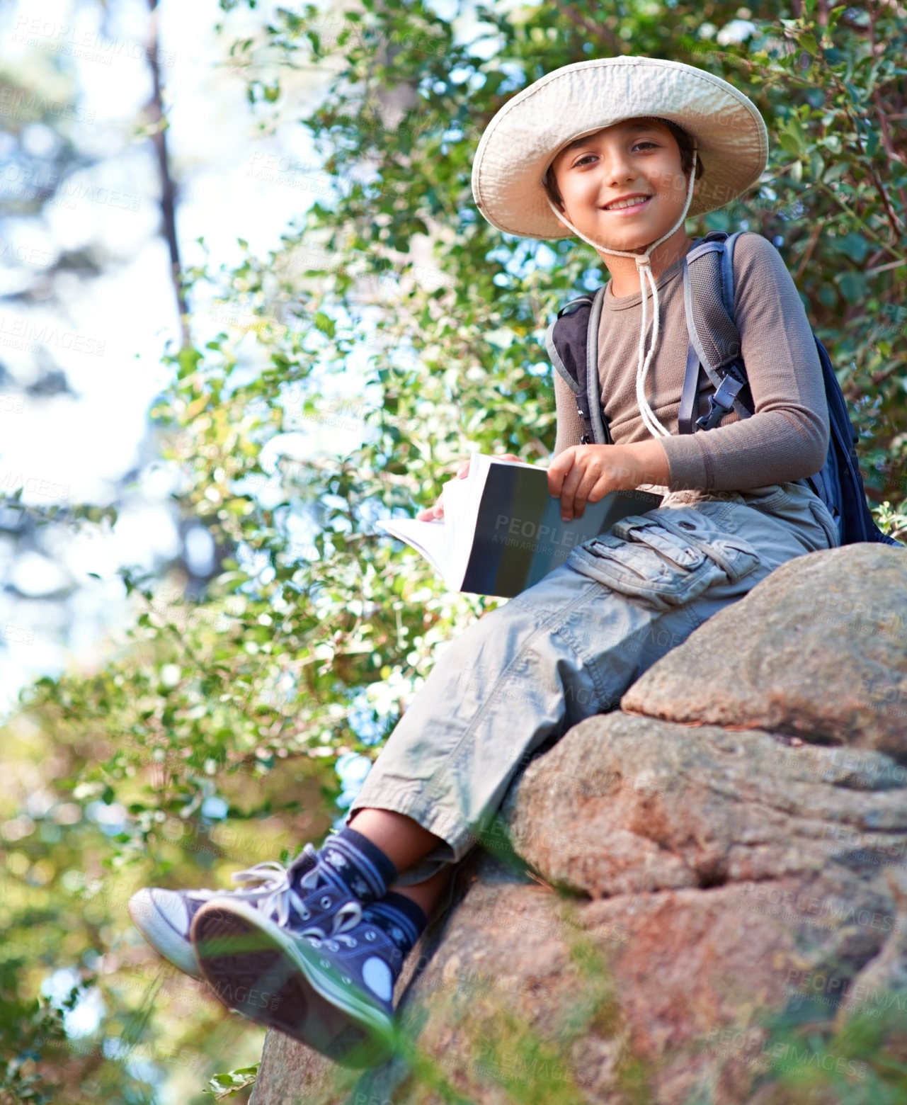 Buy stock photo Boy, kid and hiking by rock and book for reading, backpacking and adventure outdoor. Student on school field trip, relax in nature and forest for discovery or exploration, smile and travel portrait