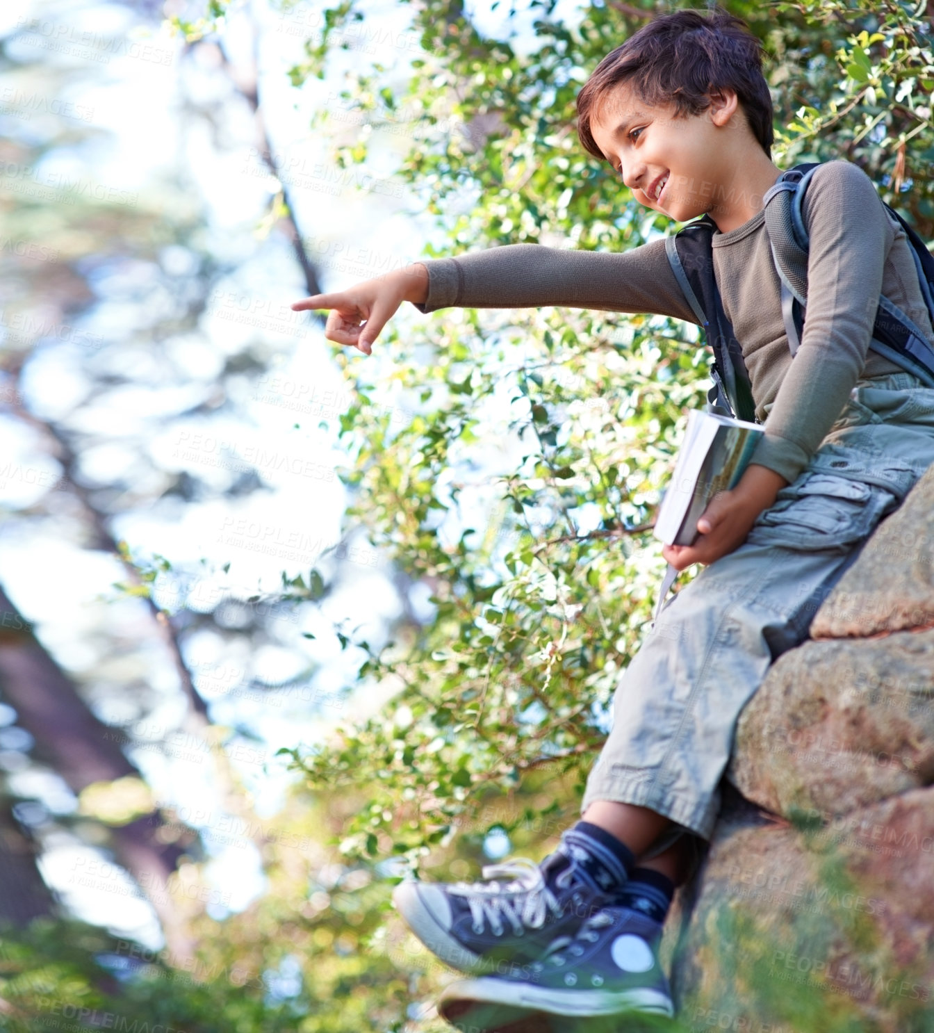 Buy stock photo A little boy with a book in his hand sitting on a rock and pointing towards something