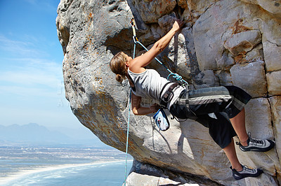 Buy stock photo A young woman climbing up a rock face while framed against a blue sky